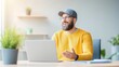 © อาริษา วันดี - Happy man in yellow sweater and cap sitting at desk with laptop, smiling and engaging during virtual meeting in bright office space with plants.