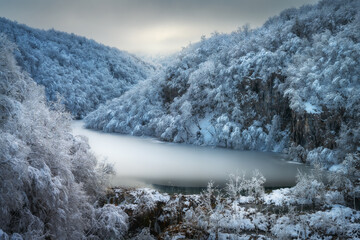 Wall Mural - Scenic view of frozen ice lake surrounded by snow-covered trees at sunset.  Landscape with lake, hills with snowy trees. Magical winter wonderland at dusk. Winter fairytale in Plitvice Lakes, Croatia
