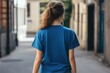 © Aleksander - A young woman walking away down a narrow street, wearing a blue color T-shirt mockupt. The scene captures a sense of solitude and exploration in an urban environment.