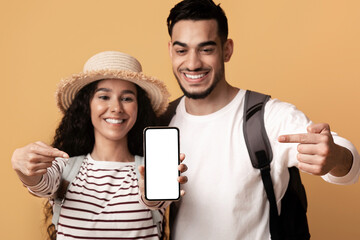 Wall Mural - Travel Booking Online. Happy Arab Travellers Couple Pointing At Smartphone With Blank Screen, Middle Eastern Spouses Recommending Mobile App For Tour Search, Standing On Yellow Background, Mockup