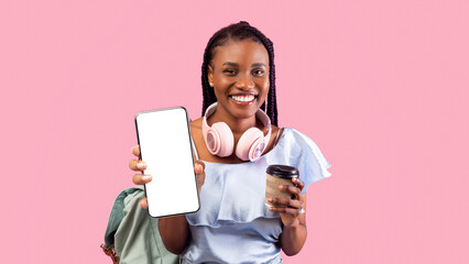 Happy black female student with backpack, headphones and coffee showing smartphone with empty screen on pink studio background, mockup for app or website design. Back to school concept