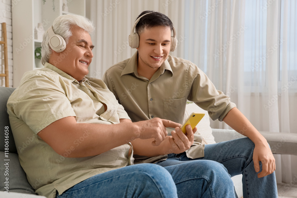 Young man with his father in headphones using mobile phone on sofa at home