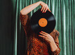 © dvulikaia - Woman in vintage leopard print dress cover her face with vinyl record on green curtain background