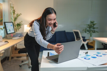 Wall Mural - Young, happy asian businesswoman talking on a mobile phone while showcasing her laptop to a colleague, working late in the office at night, immersed in collaboration and technology