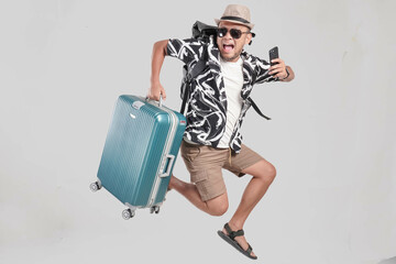 Wall Mural - Excited young Asian man tourist jumping while bringing suitcase and smartphone.