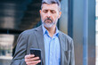 © Raquel - Smiling middle-aged businessman in a blue shirt holding and using a smartphone outdoors in a modern urban setting.