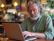 © Rodica - Mature man with beard working on laptop in cozy coffee shop environment. Elderly man using laptop in cozy café ambience for work or leisure