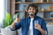 © Liubomir - Portrait of a young man with a glass of water in his hands, sitting at home on a sofa in the living room of an apartment, smiling and looking at the camera, showing a joyful thumbs up gesture.
