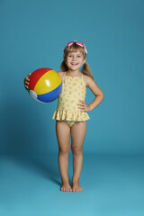 Wall Mural - Cute little girl in swimsuit with inflatable ball and swim goggles on light blue background