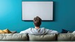 © The 2R Artificiality - Relaxed man in casual attire viewing blank television screen while seated on plush green sofa against vibrant blue wall, warm cushions in background.