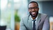 © The 2R Artificiality - Young African American businessman in gray suit smiling confidently in modern office with natural light and blurred background, showcasing professional atmosphere.