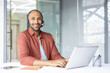 © Liubomir - Portrait of young online customer support worker. Smiling man looking at camera with headset, using laptop at workplace for video call and support consultation.