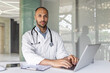 © Liubomir - Portrait experienced doctor inside a clinic office at workplace, man is looking at the camera seriously and focused, working with a laptop, preparing an electronic report of the patient's treatment.