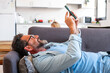 © simona - Man lying on the sofa at home in a relaxed position, using his phone to send messages to friends, scrolling through entertaining media content and videos while enjoying his leisure time