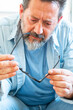 © simona - A middle-aged man sitting alone on a couch in his home, thoughtfully checking his eyeglasses, reflecting on eyesight problems and middle-age vision correction needs