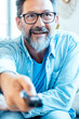 © simona - A happy adult man sitting cheerful  on a sofa in front of the television, holding a remote control, watching and changing channels, smiling and enjoying their leisure time at home