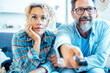 © simona - A happy adult couple sitting closely together on a sofa in front of the television, holding a remote control, watching and changing channels, smiling and enjoying their leisure time at home
