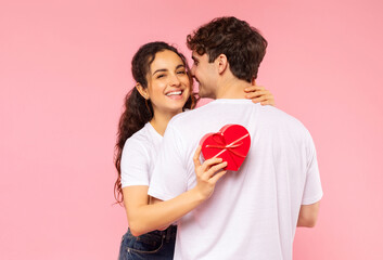 Happy couple in love celebrating Valentine's day, lady holding heart shaped gift box while embracing boyfriend, pink studio background