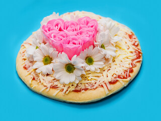 Pink roses in heart love shape and white chamomile flowers on top of a pizza and on blue background. Abstract conceptual art. Selective focus.