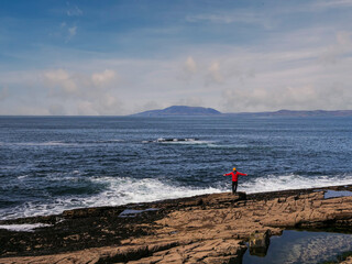 Wall Mural - A man in a red jacket stands on a rocky beach looking out at the ocean. The sky is clear and the water is calm. Mullaghmore peninsula Ireland. Travel and tourism. Trip to Irish nature