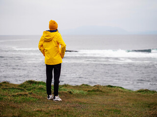 Wall Mural - A woman in a yellow jacket stands on a grassy hill overlooking the ocean. The scene is peaceful and serene, with the woman's hat adding a touch of warmth and color to the landscape. Travel and tourism