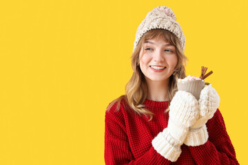 Canvas Print - Pretty young woman in mittens holding cup of hot chocolate with marshmallows and cinnamon on yellow background