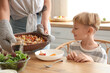 © Pixel-Shot - Young woman putting Fajita on table at dinner with her little son in kitchen