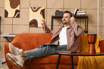 Wall Mural - Young male composer with headphones and notebook sitting on sofa in music studio