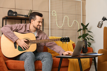 Wall Mural - Young happy male composer with laptop playing guitar on sofa in music studio