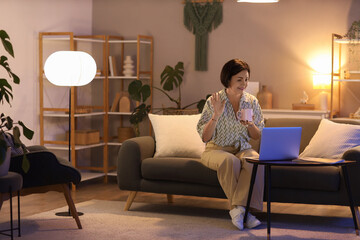 Sticker - Mature woman with cup of tea and laptop video chatting on sofa at home in evening