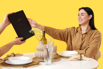 Wall Mural - Young woman taking menu from waiter at restaurant table on yellow background