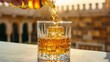 © New Stock - A hand pours golden liquid from a bottle into a crystal glass filled with ice, set against a sunlit blurred background.