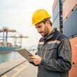 © Amina - A dock worker in a yellow helmet and gray uniform checks a tablet near stacked shipping containers