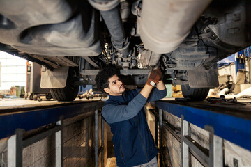 Interracial auto mechanic standing in vehicle inspection pit under chassis and repairing truck at mechanic workshop.