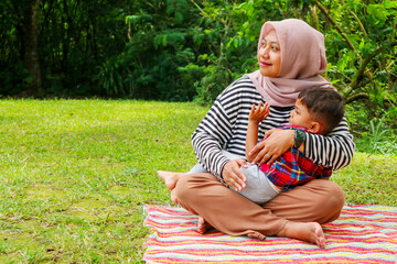 Muslim mother teaching her little son to pray with rosary beads in the park on grass field near beautiful lake. Muslim family concept.