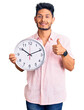 © Krakenimages.com - Handsome latin american young man holding big clock smiling happy and positive, thumb up doing excellent and approval sign