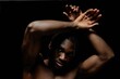 © Seventyfour - Portrait of shirtless Black man posing gracefully raising arms looking at camera with troubled eyes against black background in studio with contrast top light