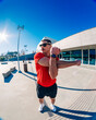 © Studio Marmellata - man doing sport in a red t-shirt and black shorts performs arm stretches under bright sunlight in a modern urban plaza with clean architectural lines and clear blue skies in the background