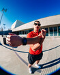 © Studio Marmellata - man doing sport in a red t-shirt and black shorts performs arm stretches under bright sunlight in a modern urban plaza with clean architectural lines and clear blue skies in the background