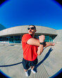 © Studio Marmellata - man doing sport in a red t-shirt and black shorts performs arm stretches under bright sunlight in a modern urban plaza with clean architectural lines and clear blue skies in the background