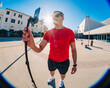 © Studio Marmellata - A man in a red t-shirt and black shorts holds a selfie stick with his smartphone attached in an urban plaza under a bright blue sky, with sunglasses shielding his eyes from the sunlight.