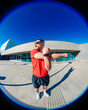 © Studio Marmellata - man doing sport in a red t-shirt and black shorts performs arm stretches under bright sunlight in a modern urban plaza with clean architectural lines and clear blue skies in the background