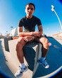 © Studio Marmellata - A young man with short curly hair sits on a concrete bench in an urban outdoor space under a bright blue sky, wearing a black t-shirt, yellow shorts, and white sneakers, with a relaxed posture