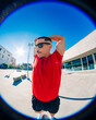 © Studio Marmellata - A man in a bright red t-shirt and black shorts stretches his arms behind his head under the bright sunlight in a spacious urban plaza, wearing sunglasses with a modern building in the background.