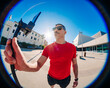 © Studio Marmellata - A man in a red t-shirt and black shorts holds a selfie stick with his smartphone attached in an urban plaza under a bright blue sky, with sunglasses shielding his eyes from the sunlight.