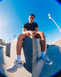 © Studio Marmellata - A young man with short curly hair sits on a concrete bench in an urban outdoor space under a bright blue sky, wearing a black t-shirt, yellow shorts, and white sneakers, with a relaxed posture