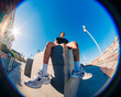 © Studio Marmellata - A young man in a black shirt and yellow shorts sits on a concrete urban structure in bright sunlight, captured with a fisheye lens against a clear blue sky and open urban space