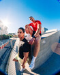 © Studio Marmellata - Two young men, one sitting on a ledge in a red shirt and the other in yellow shorts, in an outdoor urban space under a clear blue sky with a fisheye perspective capturing the setting.