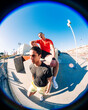 © Studio Marmellata - Two young men, one sitting on a ledge in a red shirt and the other in yellow shorts, in an outdoor urban space under a clear blue sky with a fisheye perspective capturing the setting.