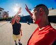 © Studio Marmellata - Two young men, one in a red shirt and one in black, exchanging a high-five outdoors with a fisheye lens capturing the vibrant sunny day and urban setting in the background.